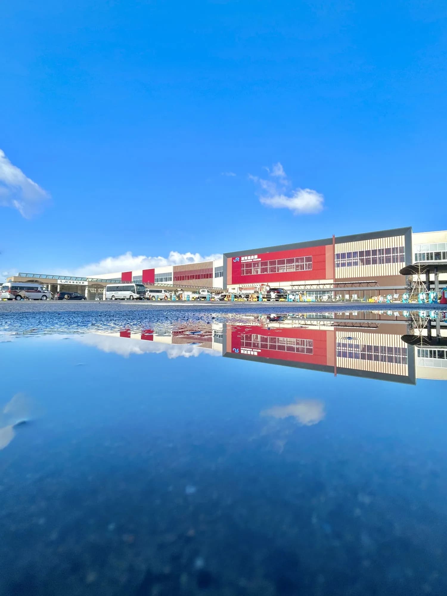 北陸新幹線 加賀温泉駅 — 青空と赤い駅舎が水たまりに映り込む